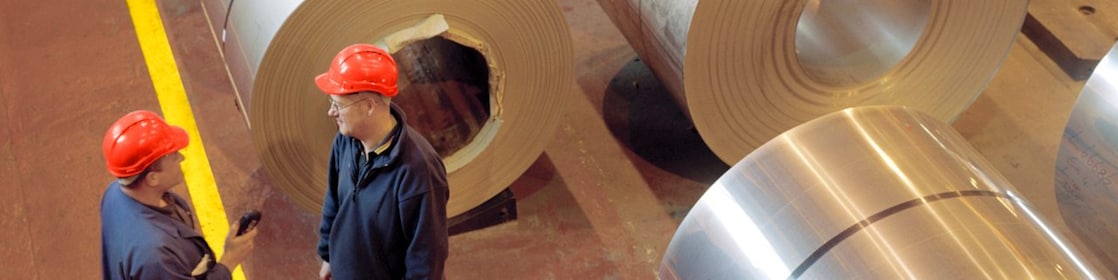 Two factory workers talking next to large rolls of steel
