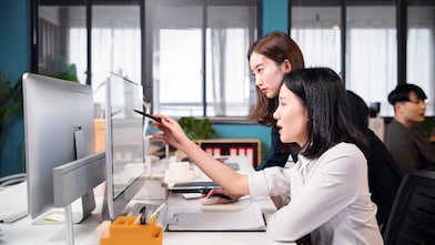 Two people collaborating at a desk, reviewing content on a computer monitor and pointing at the screen with a pen.