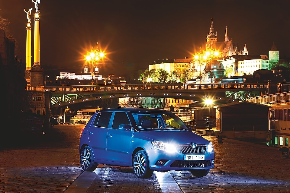 Blue car parked on a Prague road with a bridge and castle in the backdrop, at night