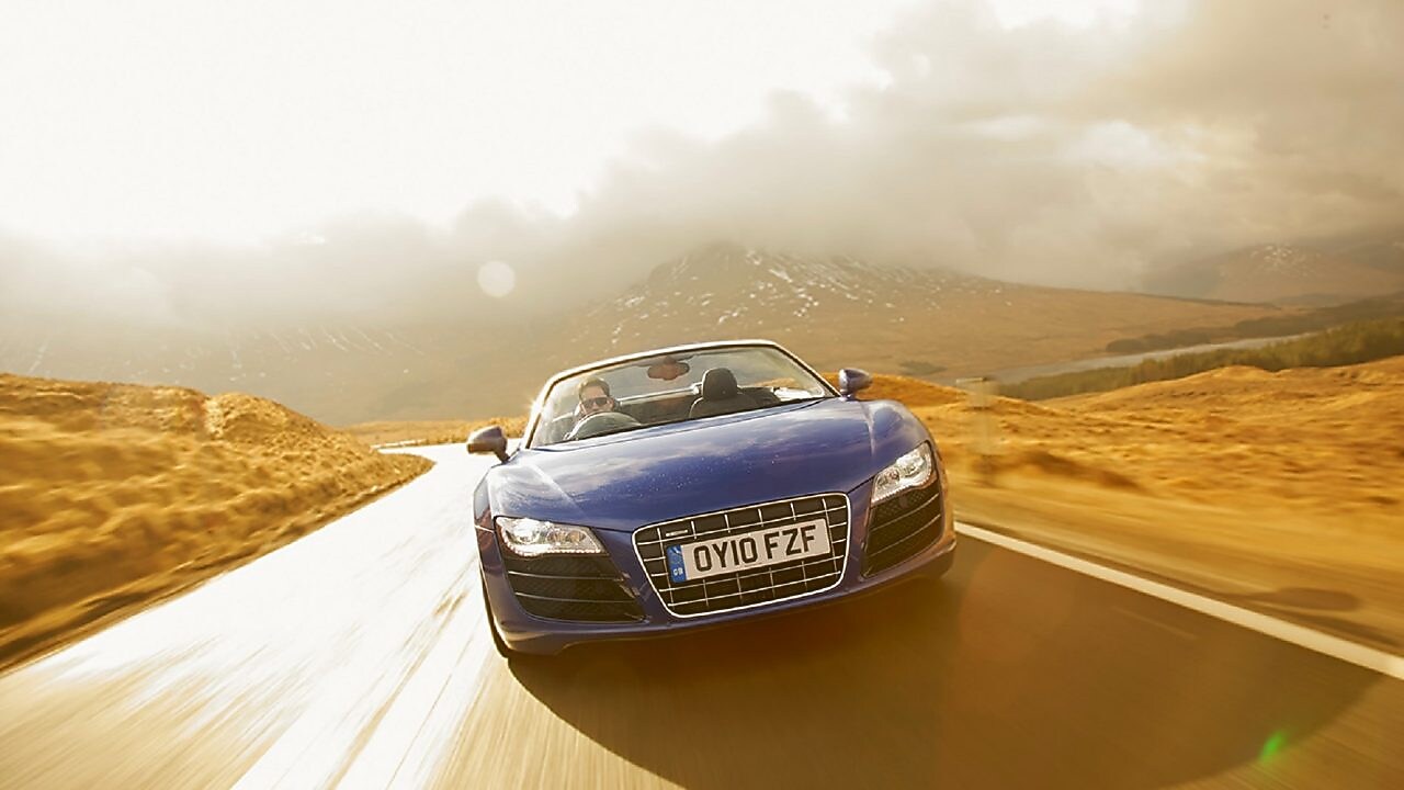  开启天窗的蓝色轿车，正面图，在被山地美景所环绕的乡间公路上行驶